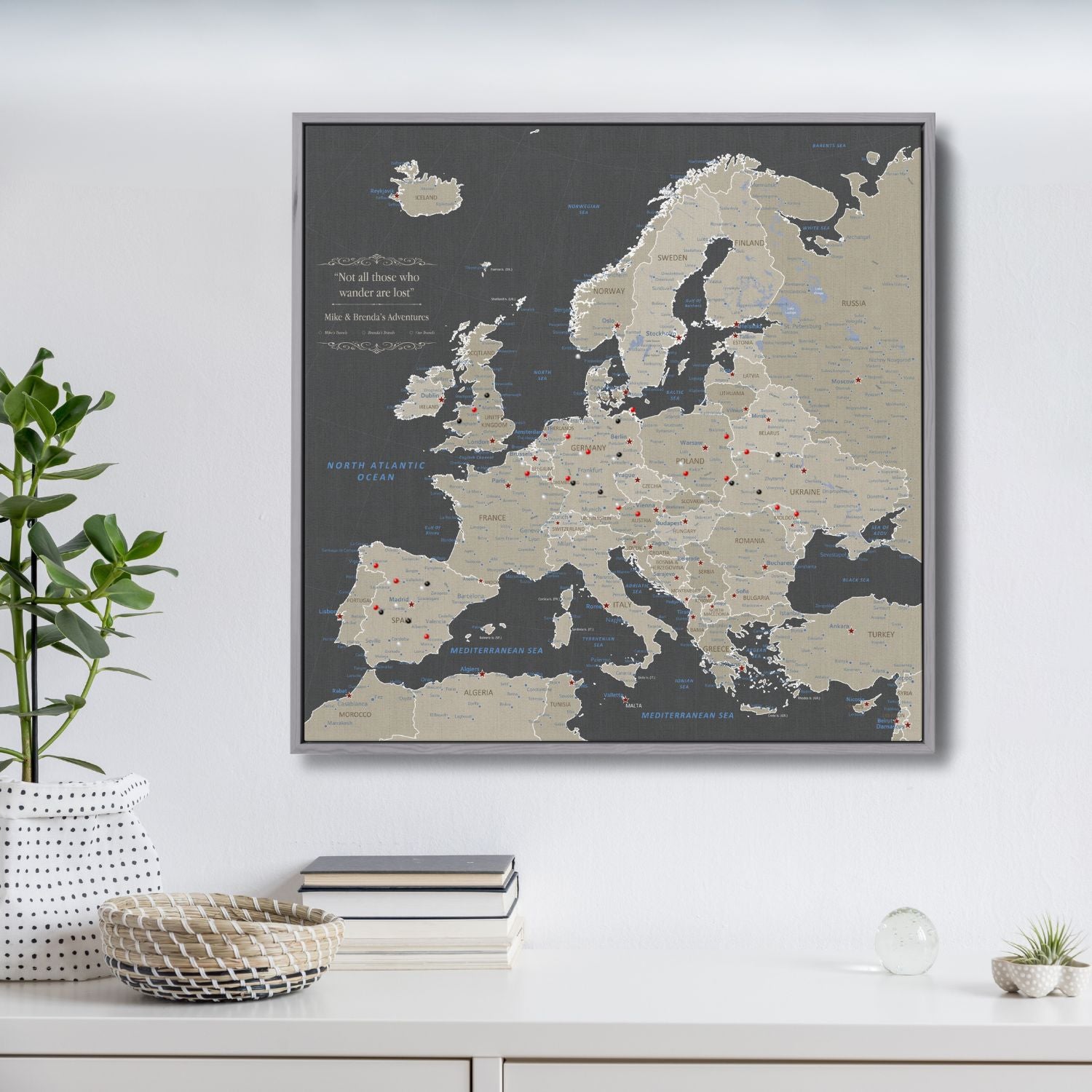 Earth toned Europe travel map canvas with red and black pins in gray float frame hanging on white wall above styled shelf with plants and books